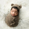 A newborn baby sleeping in a knitted bear hat and wrap set on a white fluffy surface.