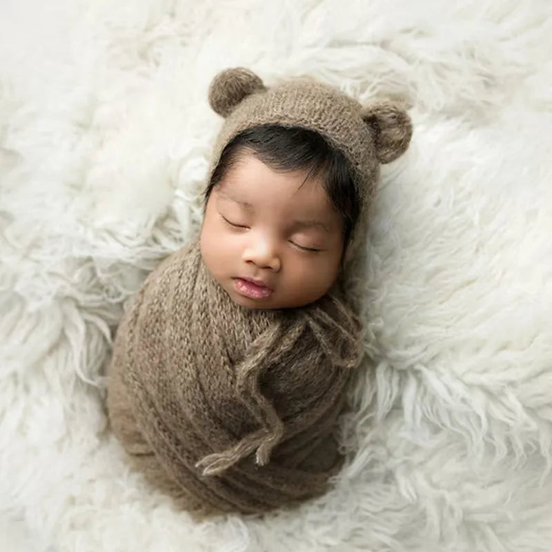 A newborn baby sleeping in a knitted bear hat and wrap set on a white fluffy surface.