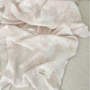 A soft pink and white muslin swaddle blanket with a delicate floral print is draped on a light-colored surface.