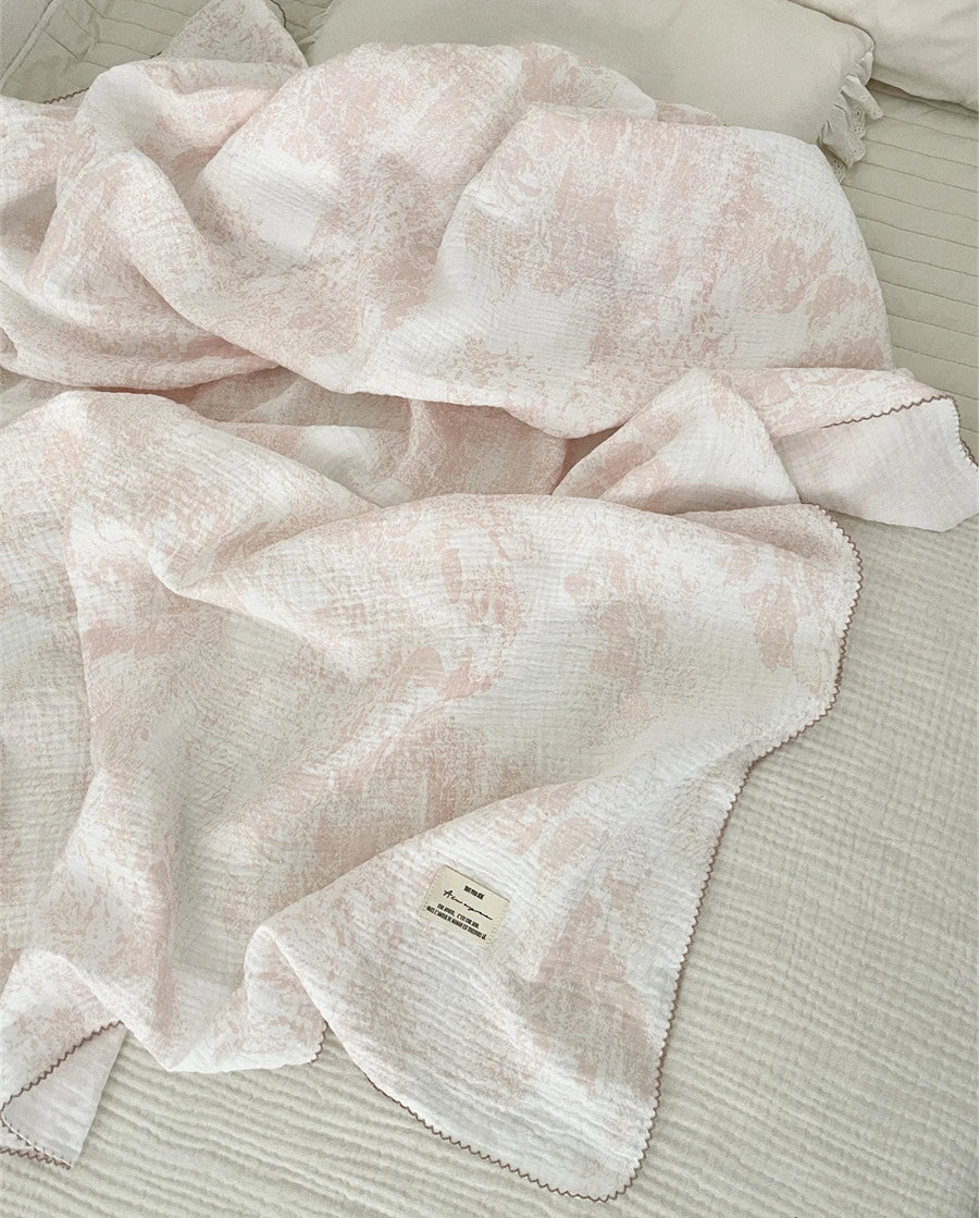 A soft pink and white muslin swaddle blanket with a delicate floral print is draped on a light-colored surface.