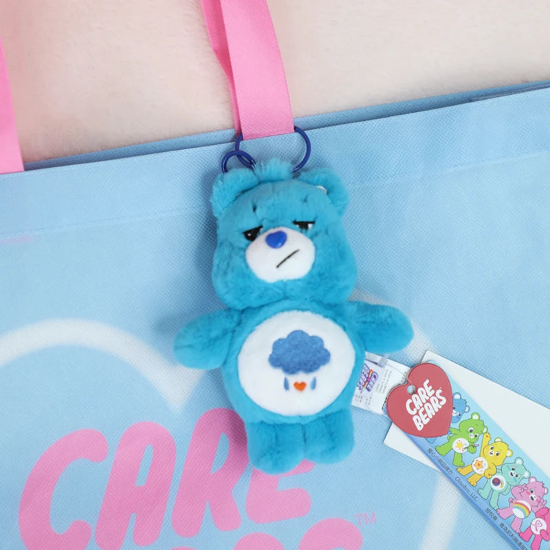 A blue Care Bears plush toy keychain charm hanging from a pink strap against a light blue fabric background.
