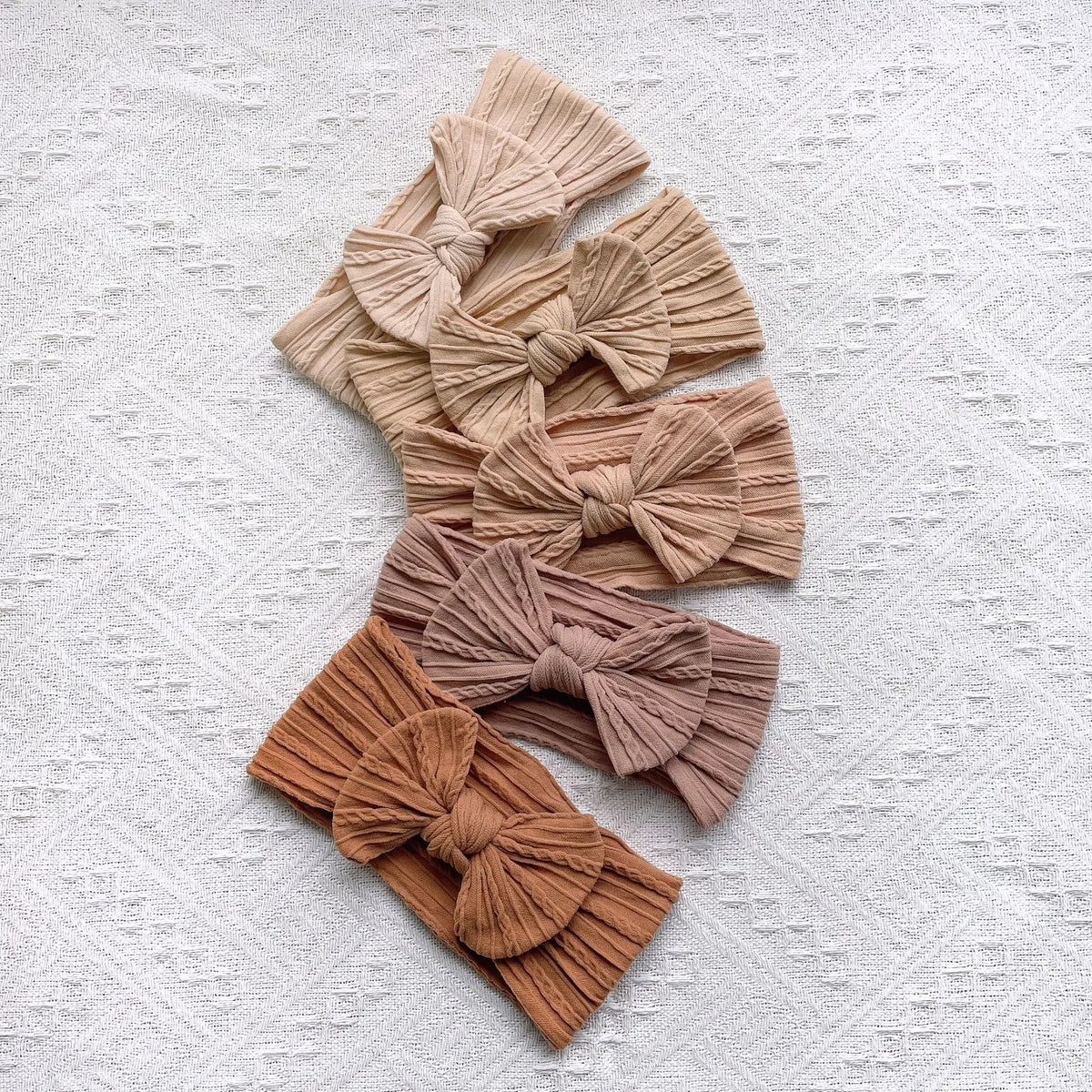 A collection of five beige and brown knitted bow baby headbands arranged in a fan shape on a white textured background.