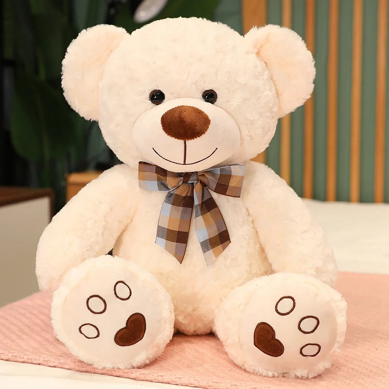 A fluffy cream-colored teddy bear plush toy with a brown nose and a plaid bow lying on a pink surface.