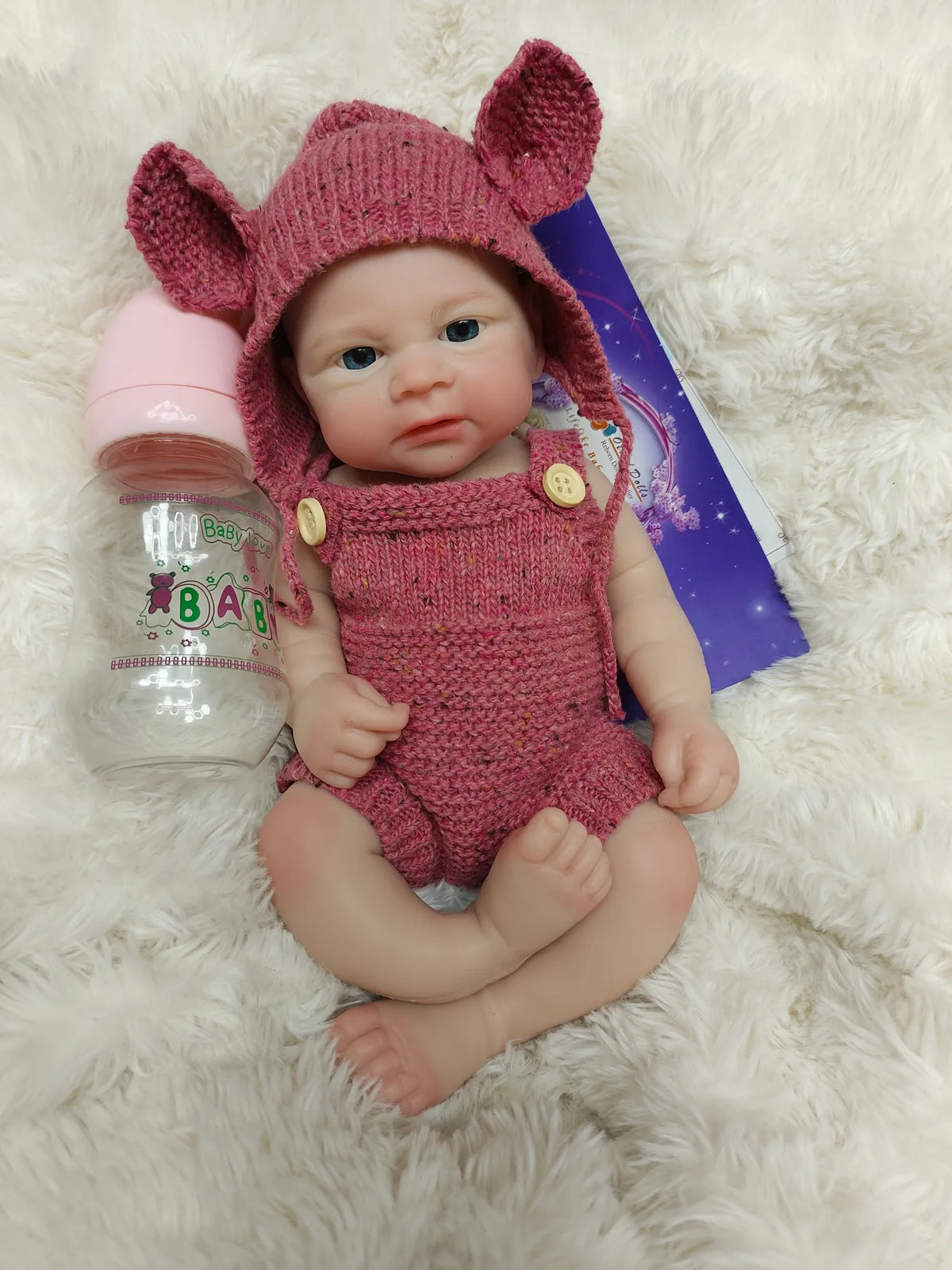 A realistic pink reborn doll with large ears is lying on a white fluffy surface next to a baby bottle.