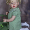 A blonde reborn baby doll with pigtails and a green dress sitting next to a green toy.