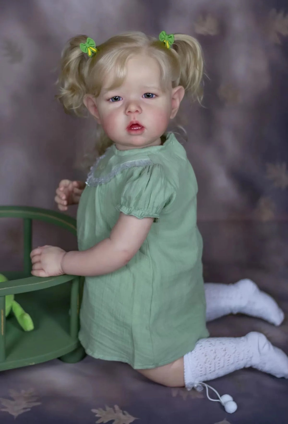 A blonde reborn baby doll with pigtails and a green dress sitting next to a green toy.
