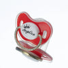 A red and clear silicone baby pacifier featuring a white center with a gold crown and the name Angelica spelled out in rhines
