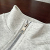 A close-up of a light gray long-sleeve sweater with a silver zipper detail on the collar.