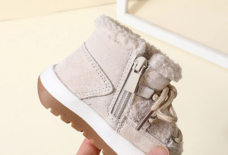 A close-up of a pair of beige baby winter snow boots with plush lining and white soles being held by a hand.