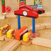 A wooden train track accessory set featuring a red station sign and an orange train on a light wood surface.