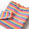 A close-up of a girls striped cotton shirt featuring rainbow accents and ruffled trim.