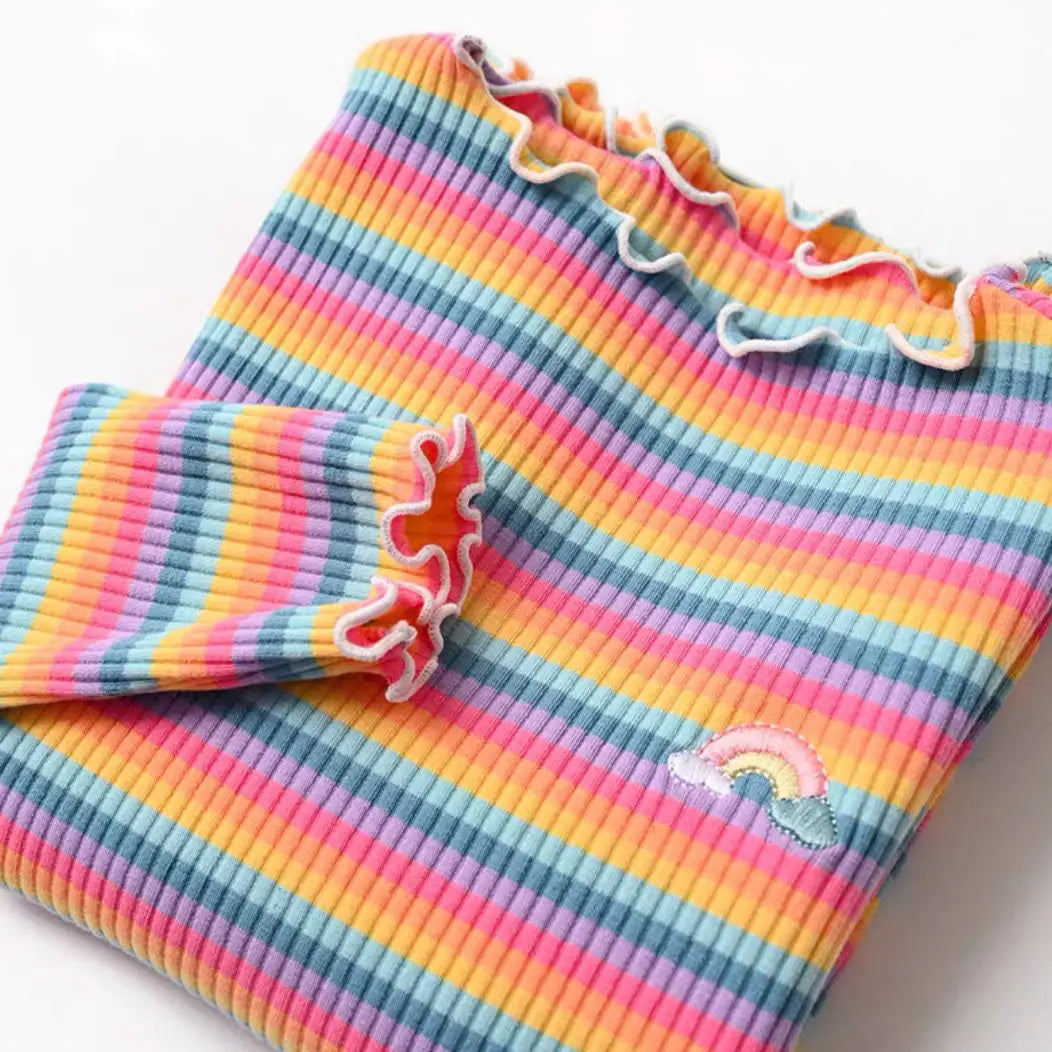 A close-up of a girls striped cotton shirt featuring rainbow accents and ruffled trim.
