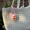 A handmade crochet mini pig keychain in pink attached to a light beige woven fabric bag.