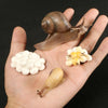 A hand holds a snail shell, a small white insect, and a small yellow insect for an animal life cycle learning set.