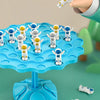 A hand is holding a white frog balance game piece above a blue plastic tree with multiple colored frog pieces.