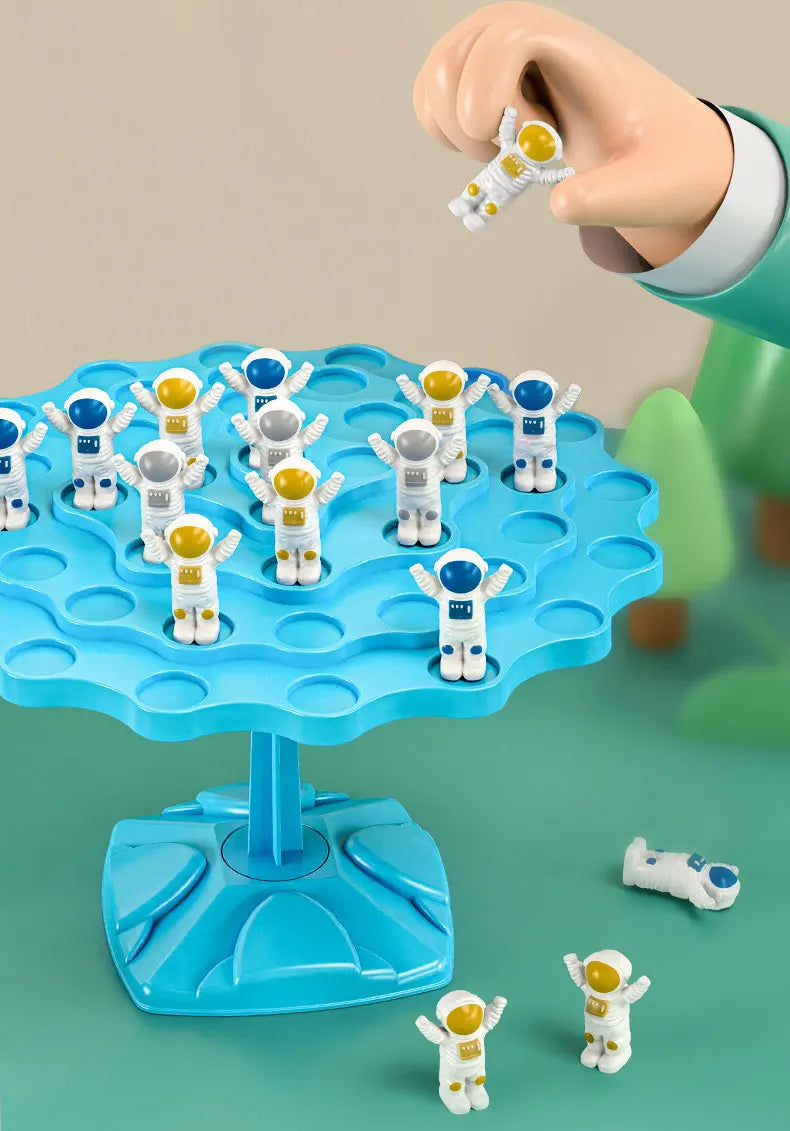 A hand is holding a white frog balance game piece above a blue plastic tree with multiple colored frog pieces.