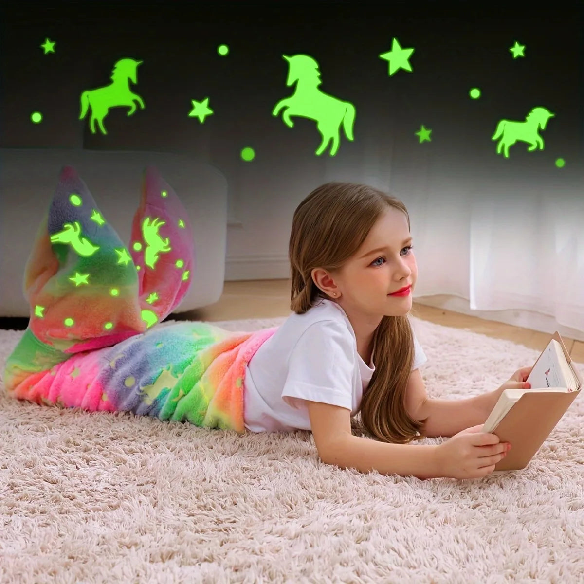 A young girl lies on a pink rug wearing a glow-in-the-dark mermaid tail blanket while looking at a book.