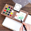 A portable wooden watercolor paint set with various colored paints and a blank canvas is shown on a wooden surface.