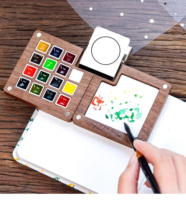 A portable wooden watercolor paint set with various colored paints and a blank canvas is shown on a wooden surface.