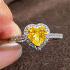 A close-up of a heart-shaped diamond ring with a yellow center stone surrounded by pave diamonds being held by a finger.
