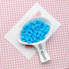A white ceramic spoon holds a pile of bright blue acrylic beads with floral edges on a pink patterned surface.
