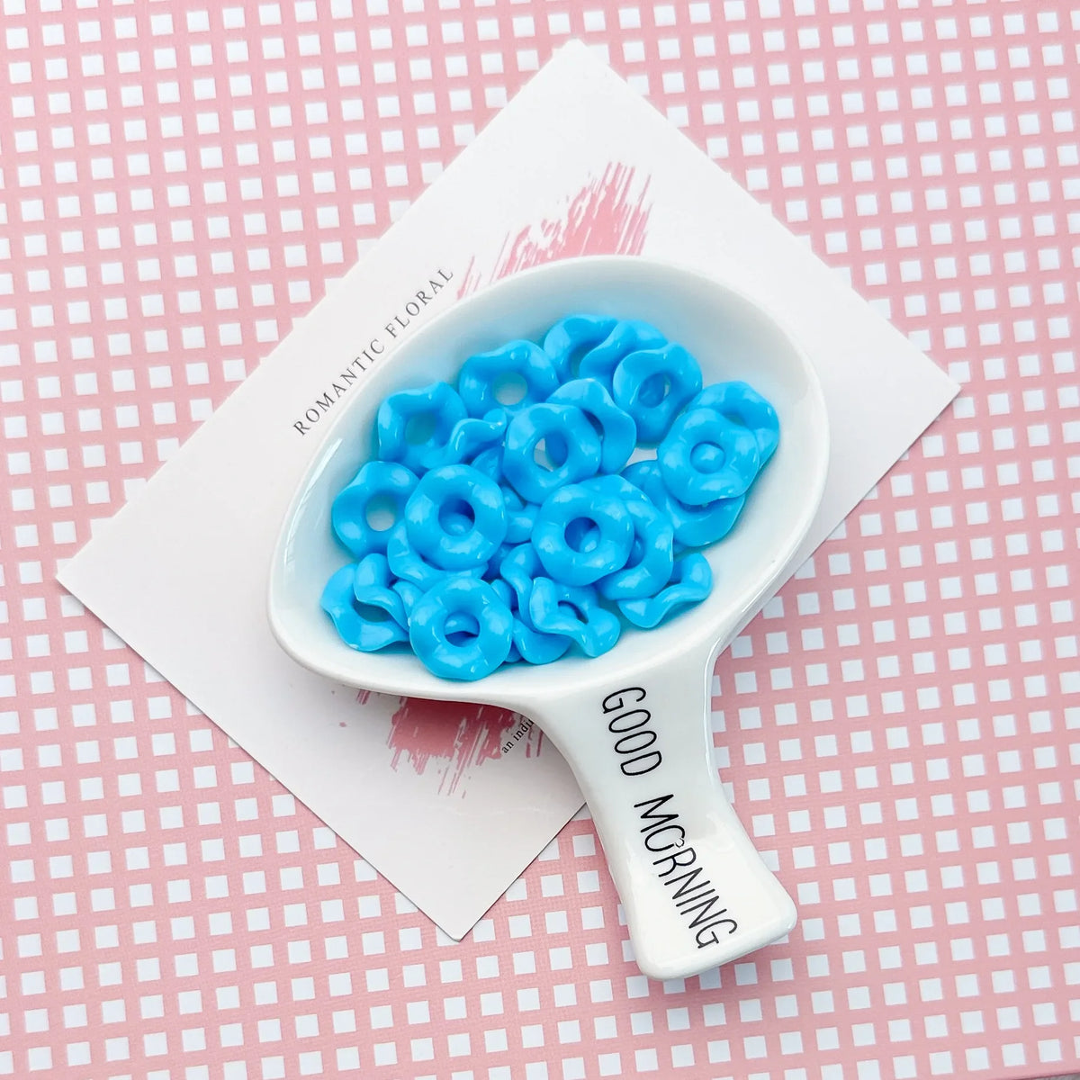 A white ceramic spoon holds a pile of bright blue acrylic beads with floral edges on a pink patterned surface.