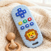 A blue silicone teether shaped like a remote control with an OK symbol and a lion face, resting on a cream-colored knitted bl