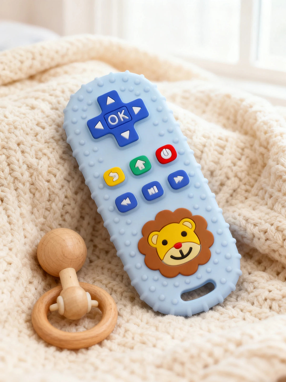 A blue silicone teether shaped like a remote control with an OK symbol and a lion face, resting on a cream-colored knitted bl