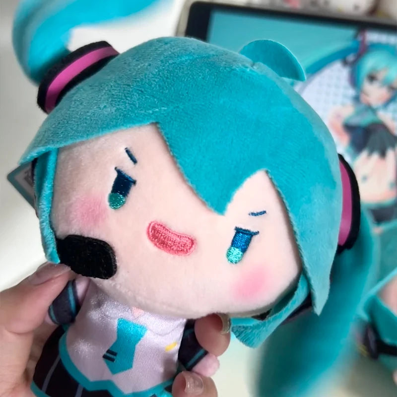 A soft teal plush doll of Hatsune Miku with pink cheeks and purple hair being held by a hand.