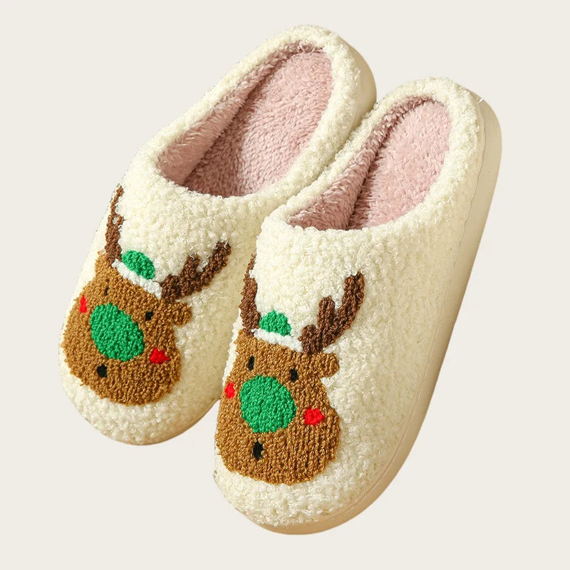 A pair of cream-colored plush indoor slippers decorated with brown reindeer faces and green accents.