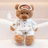 A fluffy brown bear plush toy wearing a crown and an eye mask, dressed in white pajamas.