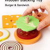 A hand is stacking wooden toy food pieces, including a green burger and sandwich piece, onto an orange base.