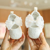 A pair of white lace-trimmed baby girl moccasin shoes with bows being held by a person's hands.