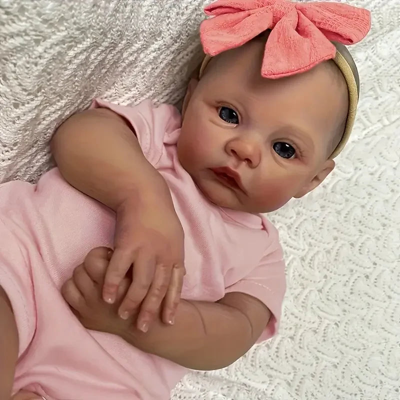A realistic reborn baby doll with light skin, blue eyes, and pink clothing, wearing a peach-colored bow.