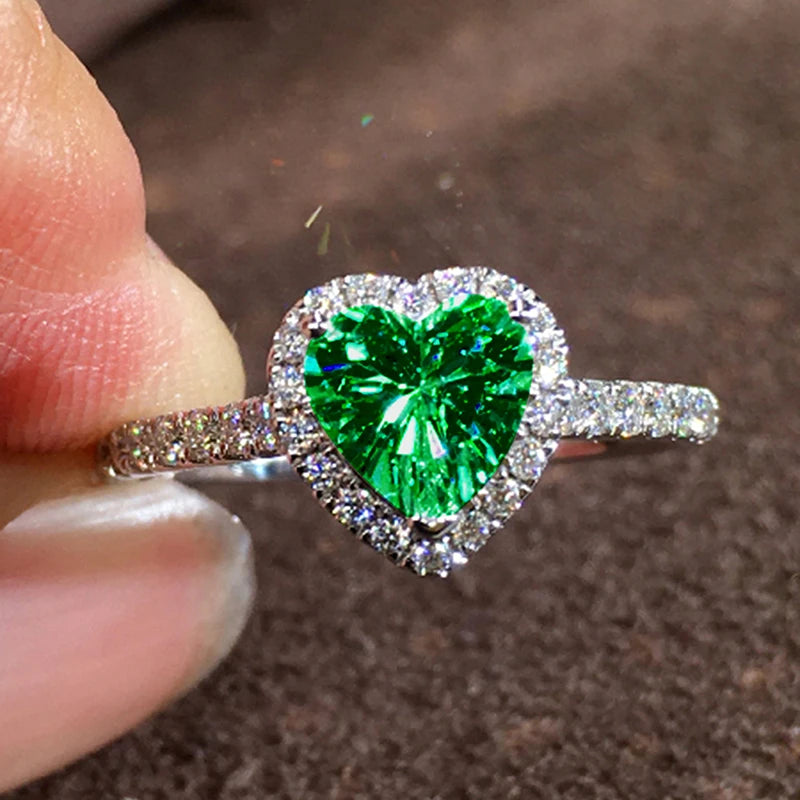 A close-up of a heart-shaped ring featuring a large, brilliant green gemstone surrounded by pave diamonds.