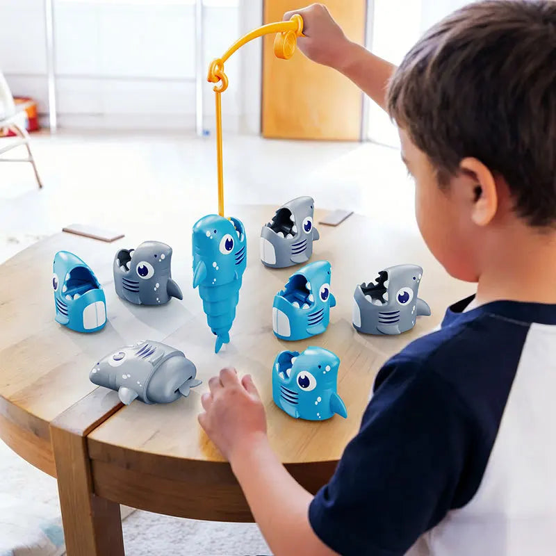 A boy is using a yellow fishing rod to retrieve eight plastic, stretchy shark figures from a wooden table.