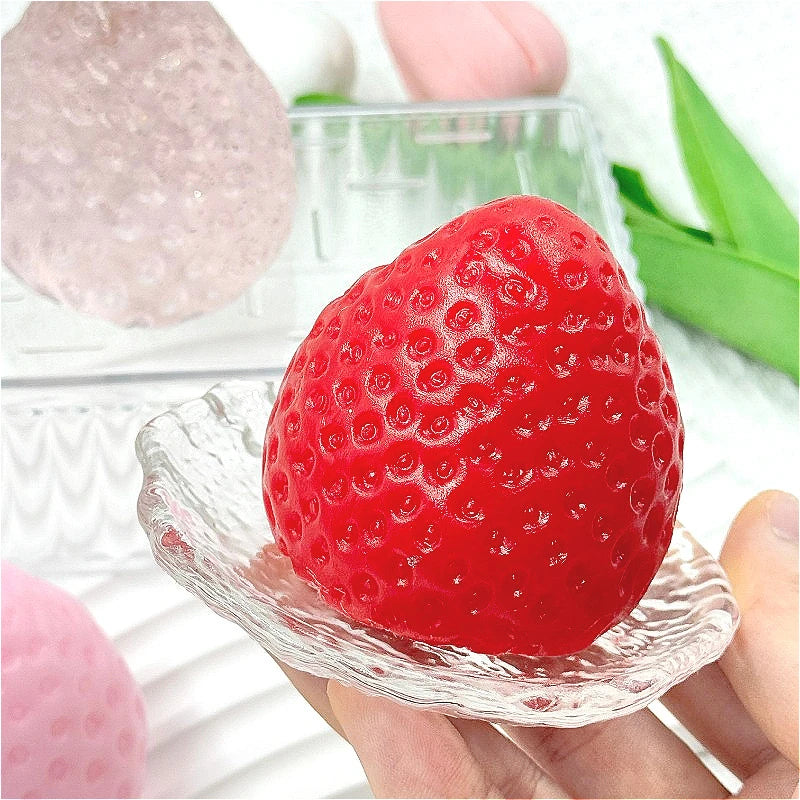 A bright red strawberry squishy toy is held in a hand, resting on a clear plastic surface.