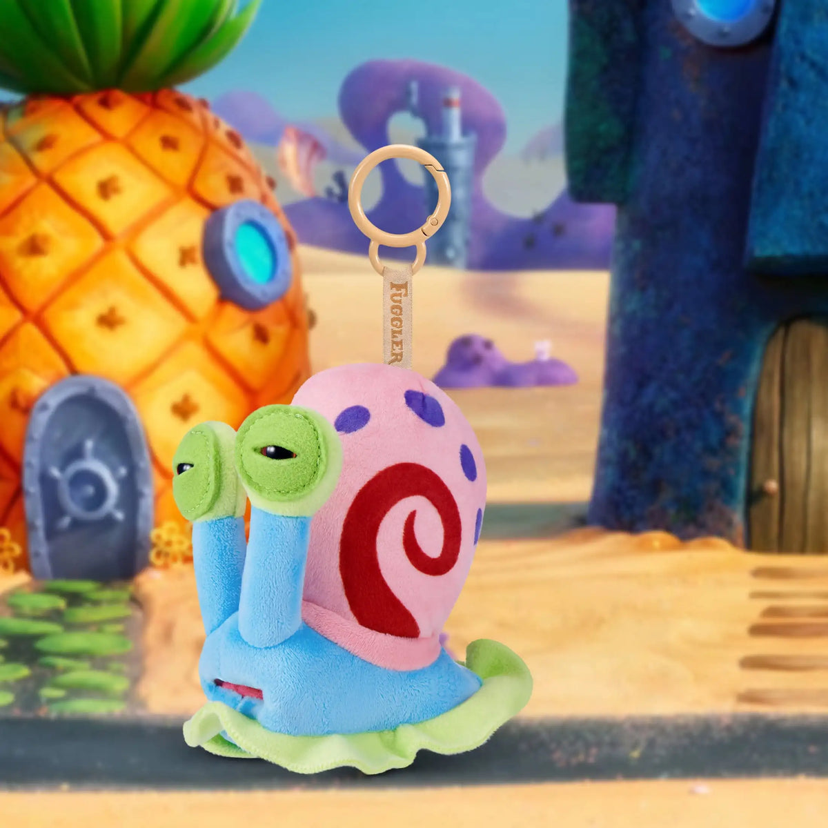 A pink and blue plush monster keychain with a spiral design is shown in a desert-like setting.
