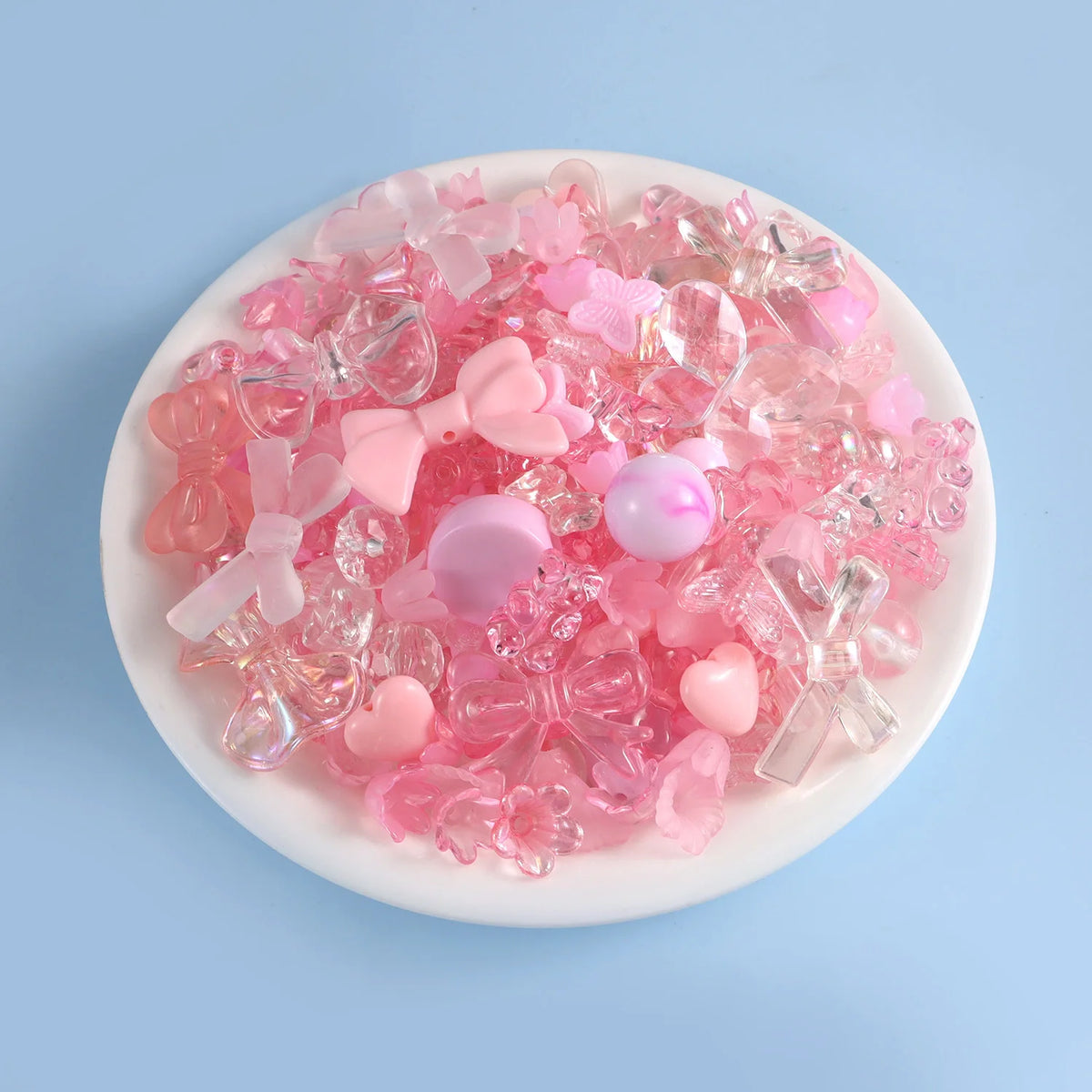 A mix of pink and clear acrylic beads, including bows and hearts, displayed in a white dish for DIY jewelry making.