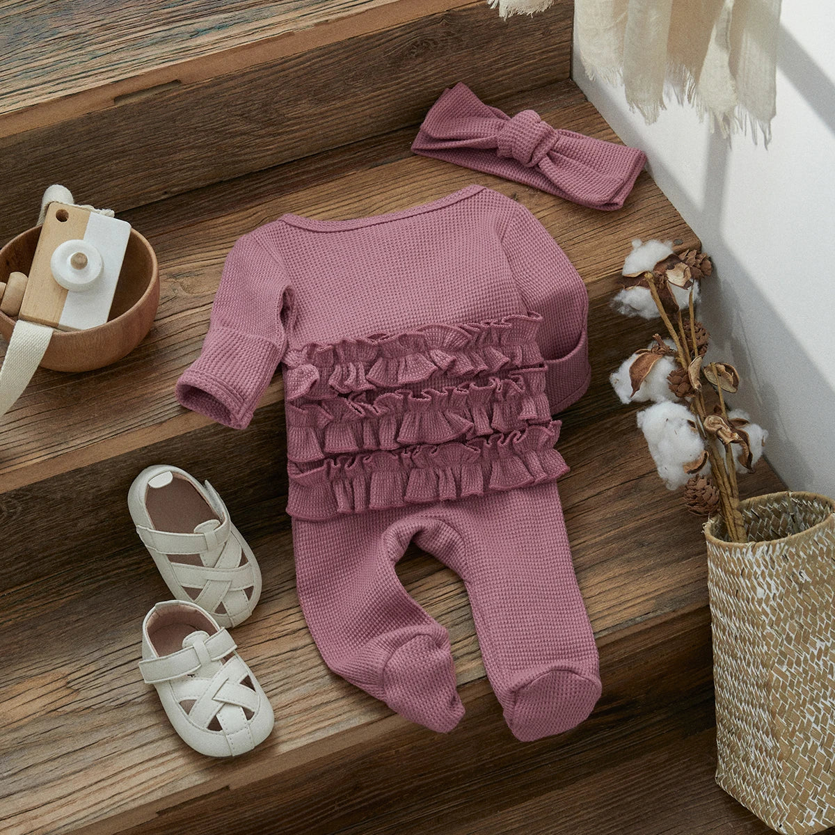 A pink knit ruffle romper for a baby girl with a matching pink bow and accessories displayed on a wooden surface.