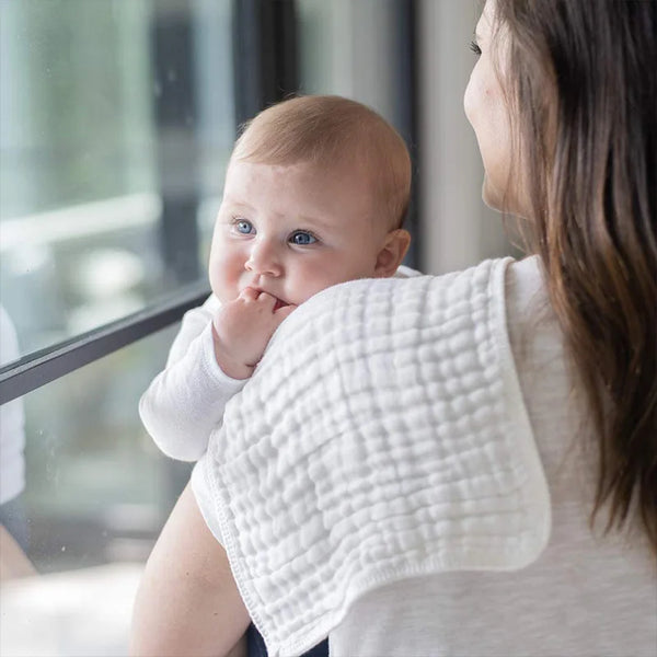 A woman holds a baby, demonstrating or using a set of three white baby burping cloths.