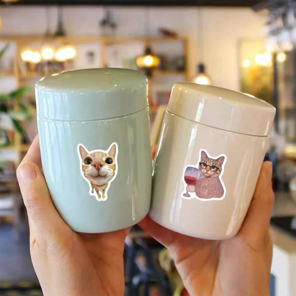 A hand holds two ceramic cups with funny cat meme stickers, one mint green and one off-white.