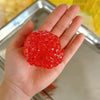 A hand holds a bright red squishy bead stress ball against a blurred background.