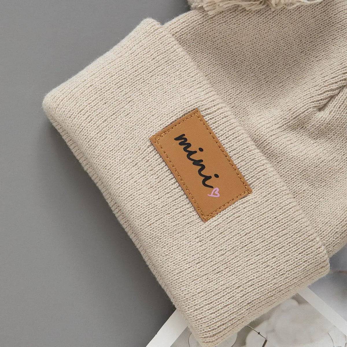 A beige knit baby beanie hat with a brown patch embroidered with the word mini