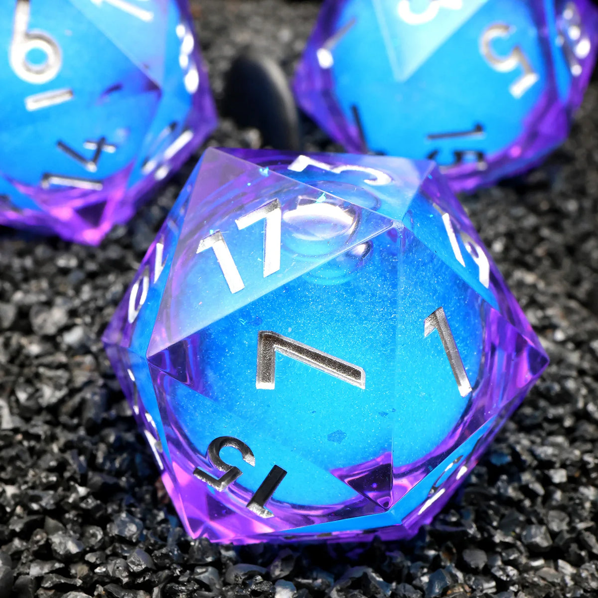 A close-up of a handmade resin D20 die with a large blue and purple liquid swirl resting on black gravel.