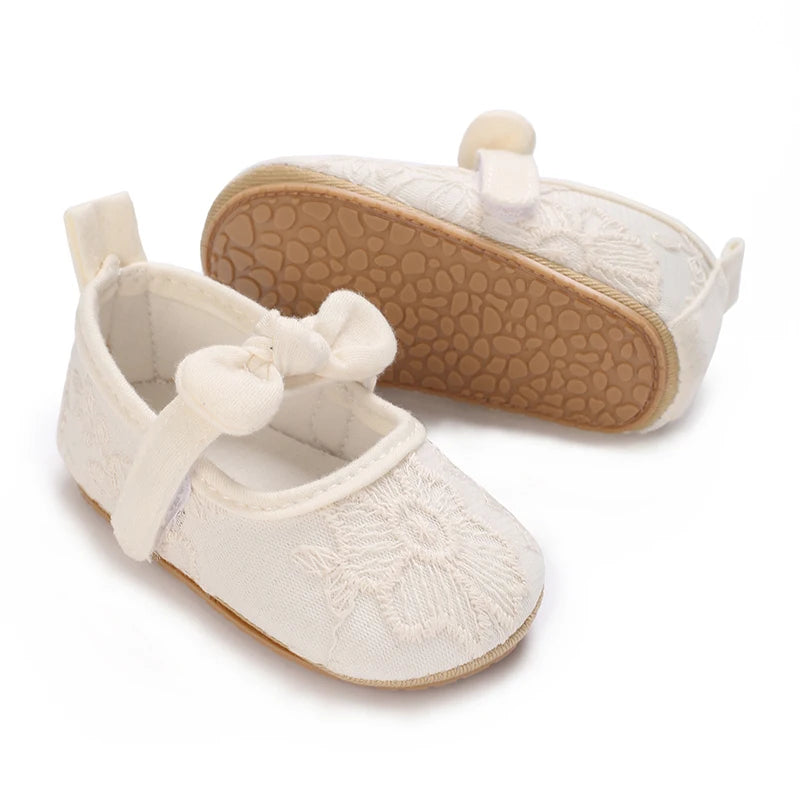 A pair of white lace-trimmed baby girl moccasin shoes with a bow and a tan sole.
