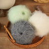 A collection of fluffy yarn balls in pale green, gray, and white resting in a woven basket.