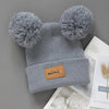 A gray knit baby beanie hat with pom-poms, featuring a small brown tag labeled mini.