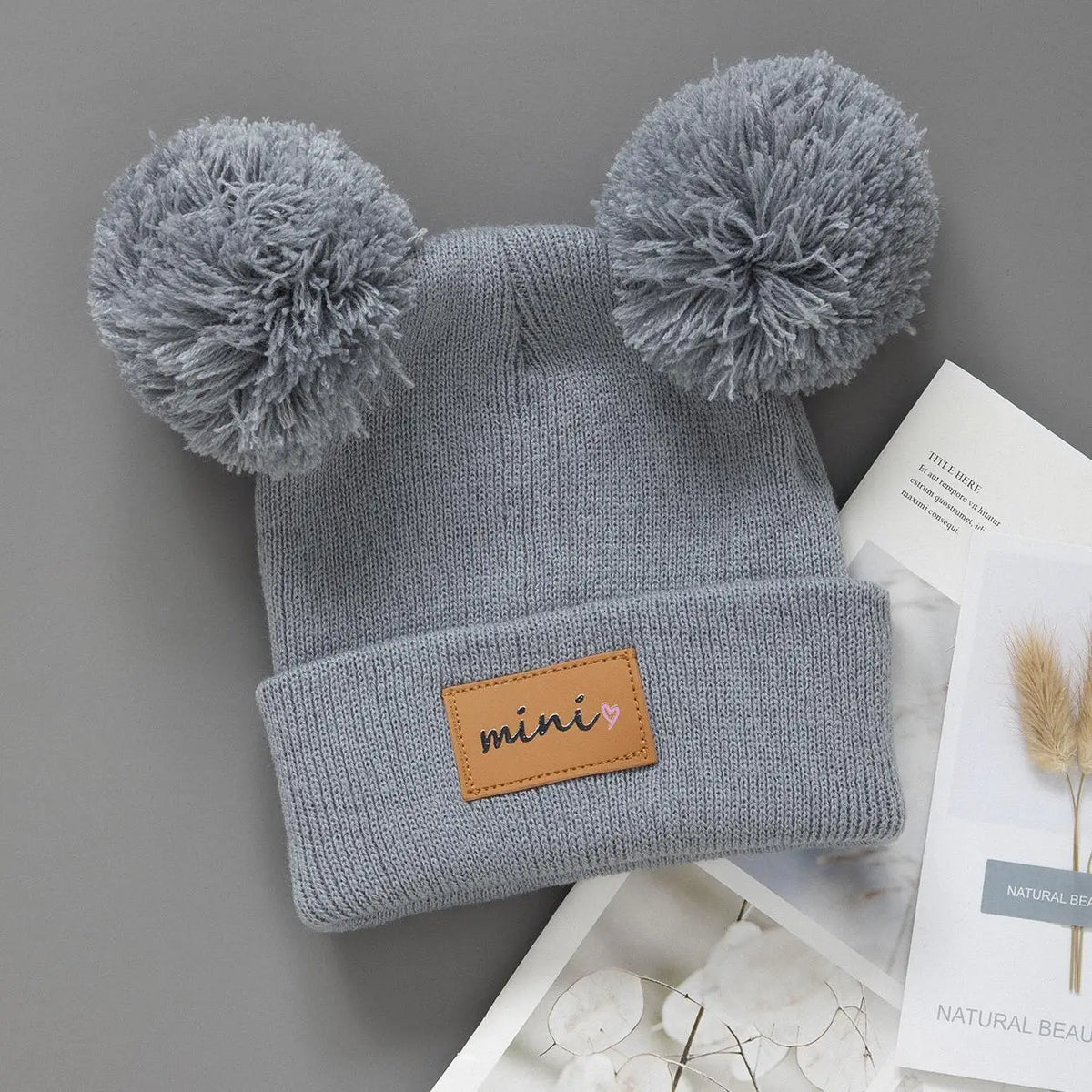 A gray knit baby beanie hat with pom-poms, featuring a small brown tag labeled mini.