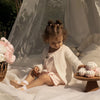 A young girl wearing pink Gladiator jelly sandals is sitting in a white fabric tent surrounded by soft light.